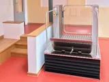 Rollstuhllift im Innenbereich neben einer Holztreppe, Plattform mit Schutzbügel und Faltenbalg